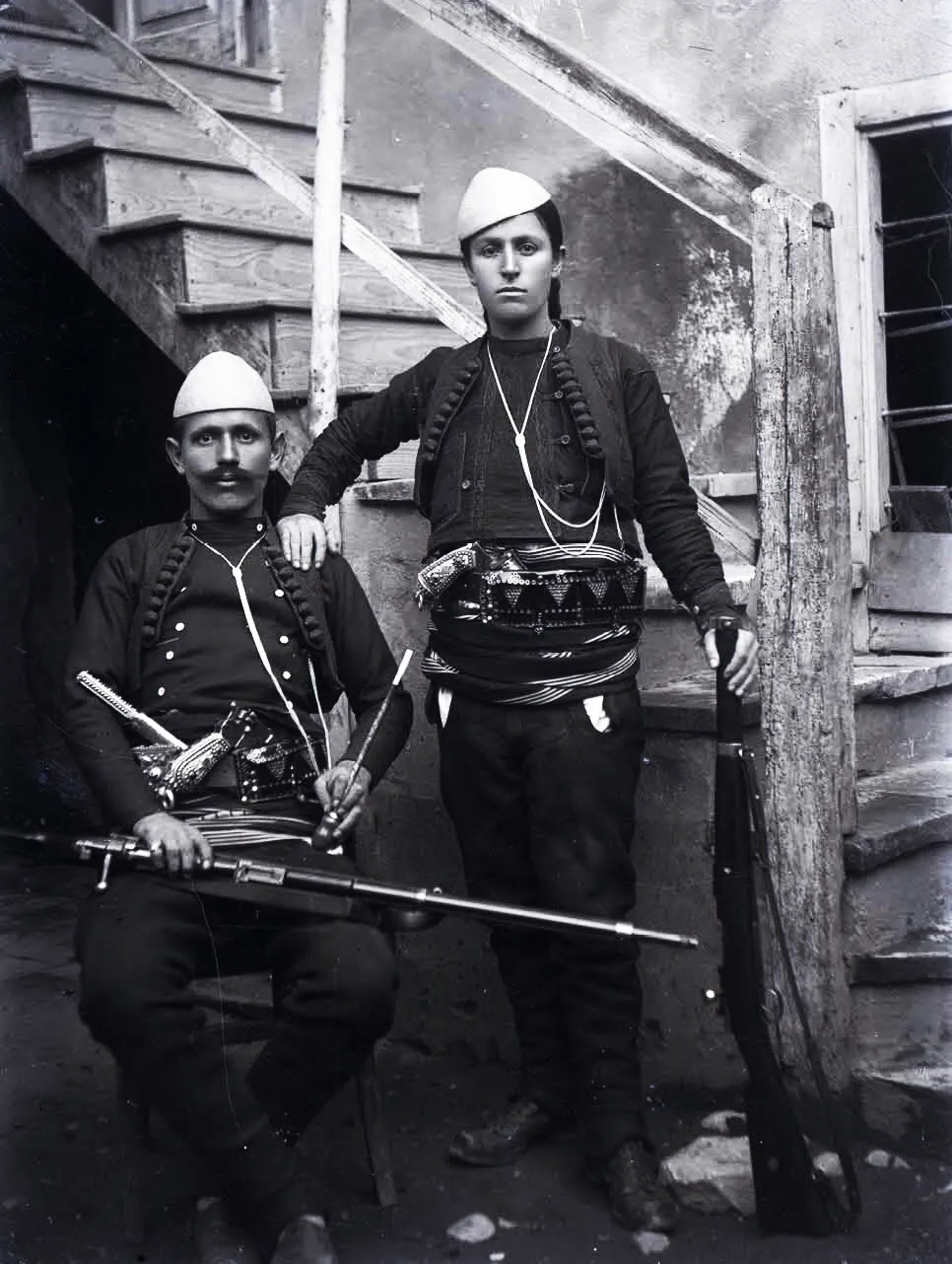 Azem Galica i ulur me pushkë dhe Shote Galica në këmbë, të armatosur, në veshje tradicionale — fotografi e Kel Marubit, rreth 1920.