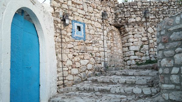 Shkallë guri dhe mure të gurta brenda kalasë së Himarës, me një portë blu në të majtë, në dritën e mbrëmjes