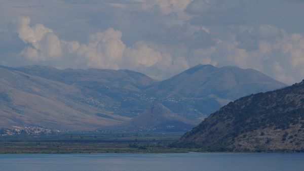 Pamje nga Korfuzi përtej ngushticës: fusha e Vrinës me kodrën e Çukës së Ajtoit dhe, në shpatet e maleve në sfond, shtëpitë e Konispolit