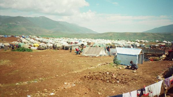 Kamp me qindra çadra të bardha të të dëbuarve kosovarë në një fushë pranë Kukësit, me male në sfond, 1999