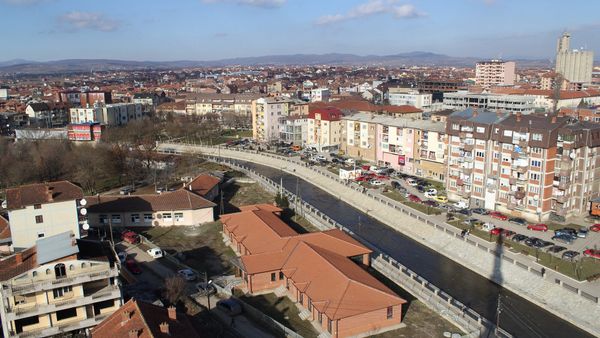 Podujeva nga lart: shtrati i kanalizuar i lumit Llap mes pallatesh e shtëpish me çati të kuqe, me kodra në horizont