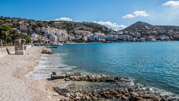 Gjiri i Sarandës me ujë të kaltër, plazhi me guralecë dhe qyteti i shtrirë mbi kodrat përreth