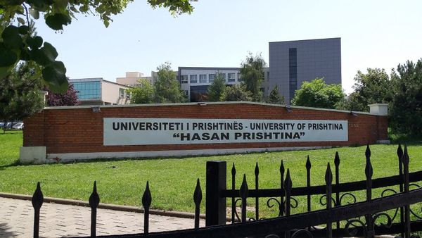 Muri me tulla në hyrje të kampusit me mbishkrimin Universiteti i Prishtinës «Hasan Prishtina», me ndërtesat e fakulteteve pas tij