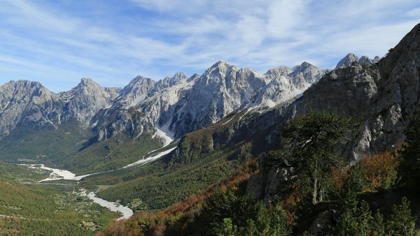 Lugina e Valbonës parë nga shpati i Qafës së Valbonës: shtrati i bardhë i lumit me zhavorr mes pyjeve në ngjyrat e vjeshtës, nën kreshtat gëlqerore të Alpeve