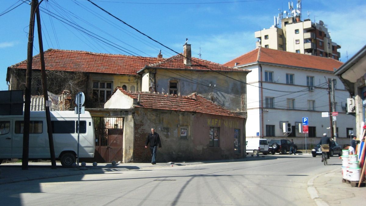 Rrugë në qendër të Bujanocit me një shtëpi të vjetër me çati me tjegulla të kuqe dhe një pallat shumëkatësh pas saj, nën qiell të kaltër