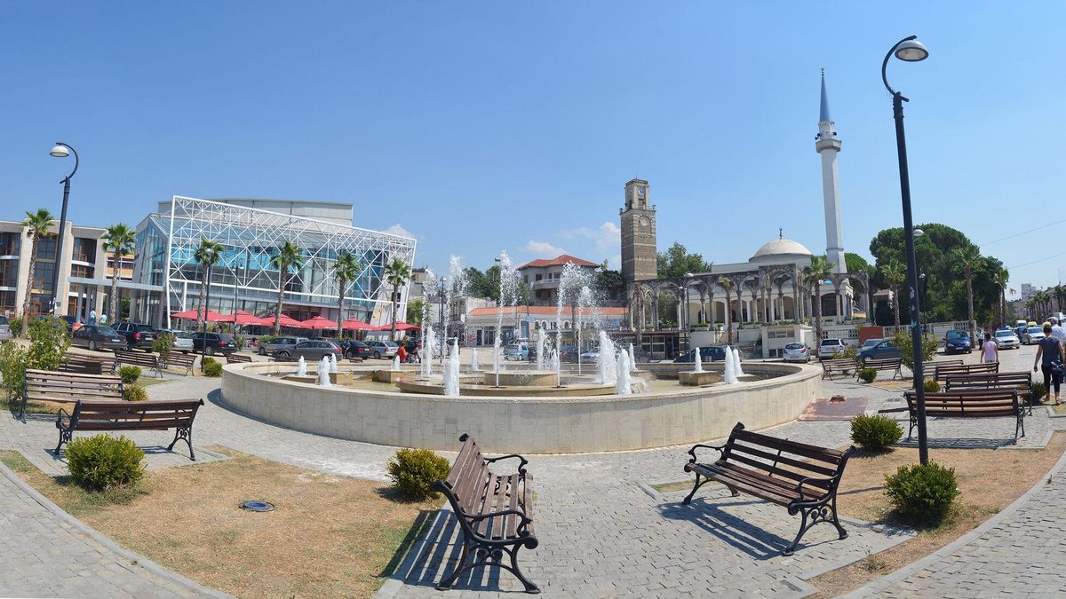 Sheshi kryesor i Kavajës me shatërvan, stola e palma, me kullën e sahatit prej guri dhe minaren e bardhë të xhamisë Kubelie në sfond