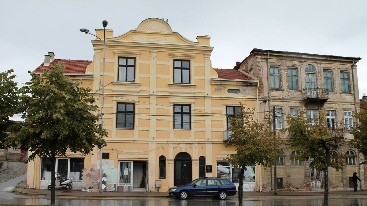Fasada e verdhë e një ndërtese dykatëshe në Manastir, me një ndërtesë tjetër të vjetër në krah dhe pemë para