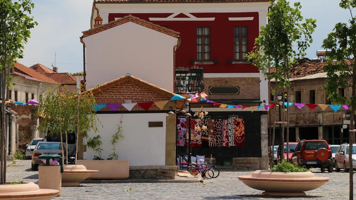 Pazari i Vjetër i Korçës: një ndërtesë dykatëshe me fasadë të kuqe, flamurka shumëngjyrëshe dhe kalldrëm në plan të parë