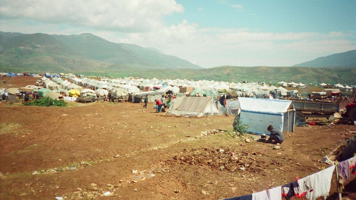 Kamp me qindra çadra të bardha të të dëbuarve kosovarë në një fushë pranë Kukësit, me male në sfond, 1999