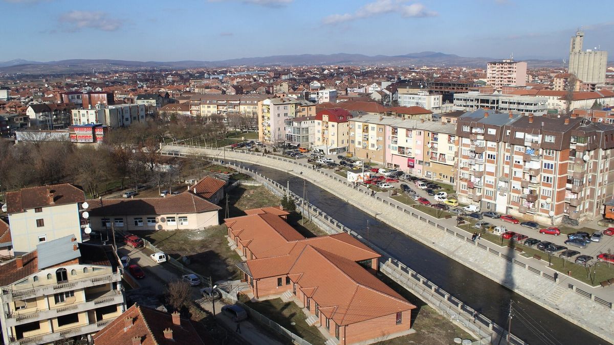 Podujeva nga lart: shtrati i kanalizuar i lumit Llap mes pallatesh e shtëpish me çati të kuqe, me kodra në horizont